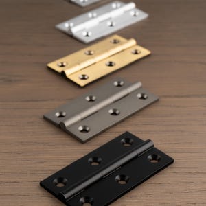 May include: A selection of door hinges in silver, gold, gunmetal, and black finishes. Each hinge is rectangular with screw holes and a central pivot, presented on a wooden surface. The hinges are for doors.