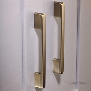 May include: Two gold-toned cabinet pulls with a rectangular shape and a slightly curved design. The pulls are mounted on a white surface.