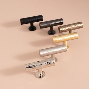 Puede incluir: Un conjunto de seis tiradores de armario de metal martillado en negro, bronce, latón, dorado y plateado. Cada tirador presenta una barra cilíndrica texturizada, montada sobre un vástago corto y una base redonda. Adecuado para armarios de cocina o baño.