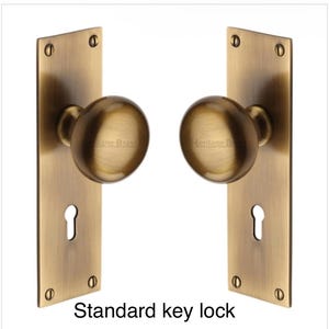 Peut inclure: Deux poign&eacute;es de porte en laiton antique avec plaques rectangulaires. Chaque plaque comporte une serrure et est fix&eacute;e avec quatre vis. Les boutons ronds portent l'inscription "Heritage Brass". Le texte "Standard key lock" est en bas.