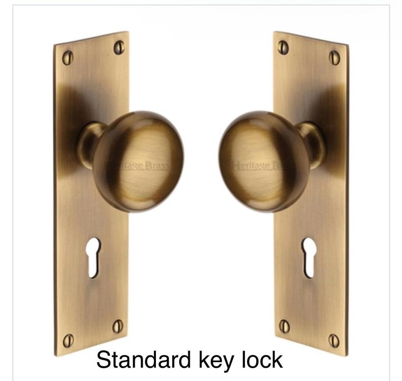 Peut inclure: Deux poign&eacute;es de porte en laiton antique avec plaques rectangulaires. Chaque plaque comporte une serrure et est fix&eacute;e avec quatre vis. Les boutons ronds portent l'inscription "Heritage Brass". Le texte "Standard key lock" est en bas.