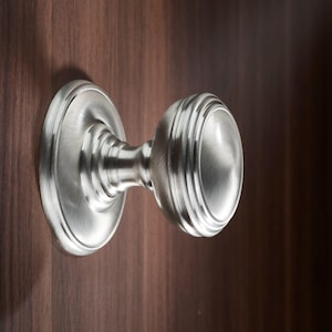 Pode incluir: Uma maçaneta de armário prateada com um design redondo e canelado. A maçaneta está montada em uma superfície de madeira.