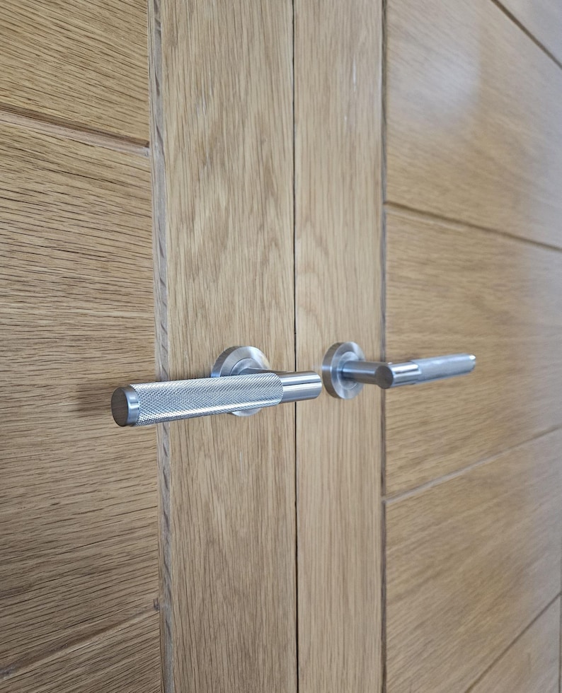May include: Close-up of a pair of wooden doors with silver metal door handles. The handles have a textured, knurled grip and a cylindrical end. The doors are made of light-colored wood with horizontal grain.