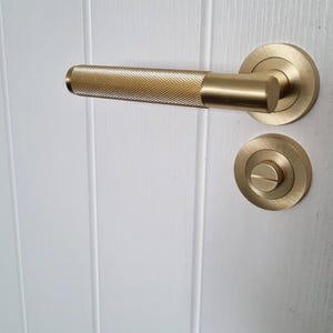May include: A gold-colored door handle and lock set. The handle has a textured, diamond-patterned grip. The round escutcheon and the handle's base are smooth. The hardware is mounted on a white, vertical-paneled door.