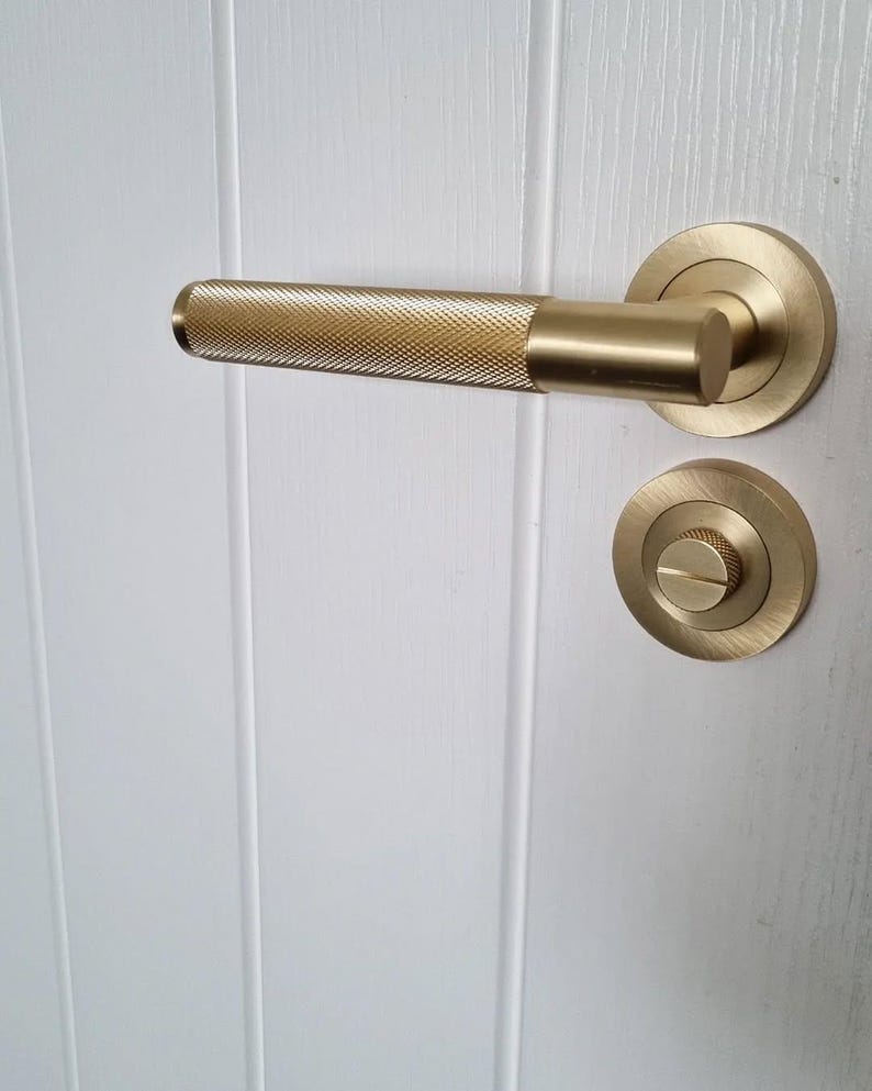 May include: A gold-colored door handle and lock set. The handle has a textured, diamond-patterned grip. The round escutcheon and the handle's base are smooth. The hardware is mounted on a white, vertical-paneled door.