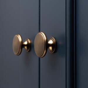 May include: Two round, gold-toned cabinet knobs with a raised edge and a slightly curved base. The knobs are mounted on a dark blue cabinet door.