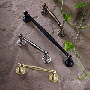May include: A collection of cabinet hardware in various finishes, including black, silver, and gold. The handles have a classic design with rounded ends and decorative bases. The image is set against a textured stone background.