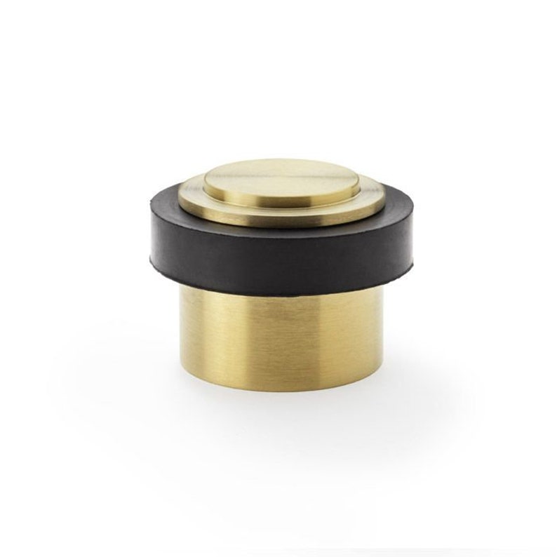 May include: A gold doorstop with a black rubber base. The doorstop has a round top and a cylindrical base.