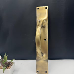 以下が含まれることがあります： ブロンズ色のドアハンドル。曲線的なハンドルと長方形のバックプレートが付いています。ハンドルはテクスチャ加工された表面で、4本のネジで固定されています。ドアハンドルはドア用の装飾要素です。