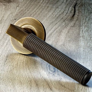 Könnte beinhalten: Ein bronzefarbener Türgriff mit einem strukturierten, zylindrischen Griff und einer kreisförmigen Rückplatte. Der Griff hat einen rechteckigen Hebel und ist auf einer hellen Holzmaserungsoberfläche angebracht. Das Design hat eine Vintage- oder Antik-Ästhetik.