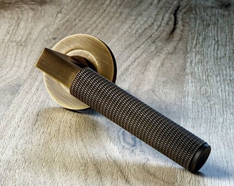 Pode incluir: Uma maçaneta de porta em tom bronze com uma pega cilíndrica texturizada e uma placa traseira circular. A maçaneta tem uma alavanca retangular e está colocada sobre uma superfície de grão de madeira clara. O design apresenta uma estética vintage ou antiga.