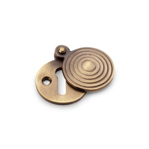 May include: A brass keyhole cover with a circular design. The cover has a concentric circle pattern and a hinged lid.