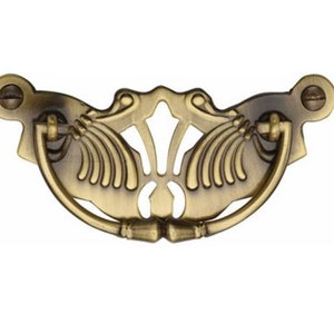 Könnte beinhalten: Ein goldfarbener, verzierter Metall-Schrankgriff. Der Griff hat eine gebogene Form und ein zentrales Element mit einem floralen Motiv.