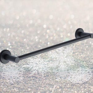 Puede incluir: Barra porta toallas de metal negro con extremos redondeados. La barra está montada sobre un fondo blanco.