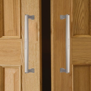 May include: Two silver door handles with a brushed finish are mounted on wooden doors. The handles are rectangular with rounded edges and have a small, curved lever on each side. The handles are labeled "Heritage Brass" in a black font.