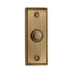 May include: A brass doorbell button with a round, raised button in the center. The button is surrounded by a rectangular frame.