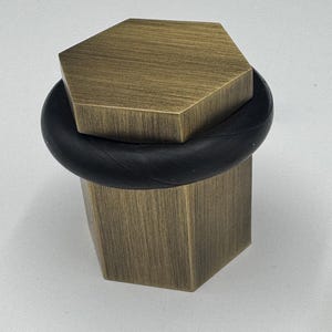 May include: A hexagonal brass doorstop with a black rubber stopper. The doorstop has a brushed finish.