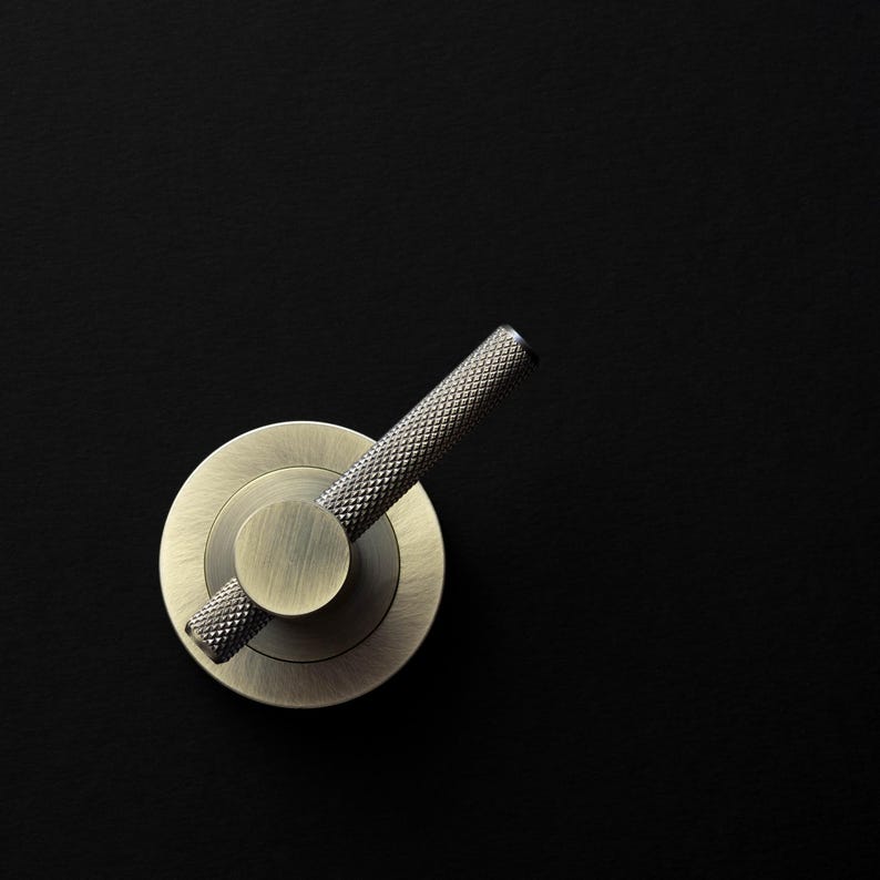 May include: A close-up of a brass-toned door handle against a black background. The handle features a circular base with a small, round center and a textured, angled lever. The metal has a vintage, antique appearance.