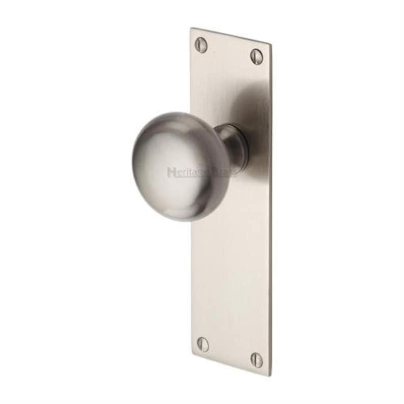 Peut inclure: Un bouton de porte en nickel bross&eacute; avec une poign&eacute;e ronde et une plaque arri&egrave;re rectangulaire. La plaque arri&egrave;re est fix&eacute;e avec quatre vis. Le texte "Heritage Brass" est visible sur le bouton.