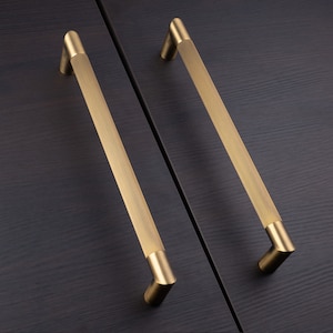 Puede incluir: Dos tiradores de armario dorados con un diseño acanalado. Los tiradores están montados en una superficie de madera oscura.