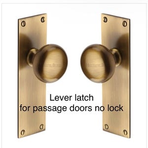 Peut inclure: Deux loquets &agrave; levier en laiton antique pour portes de passage, chacun avec un bouton rond et une plaque arri&egrave;re rectangulaire. Le texte "Lever latch for passage doors no lock" est imprim&eacute;.