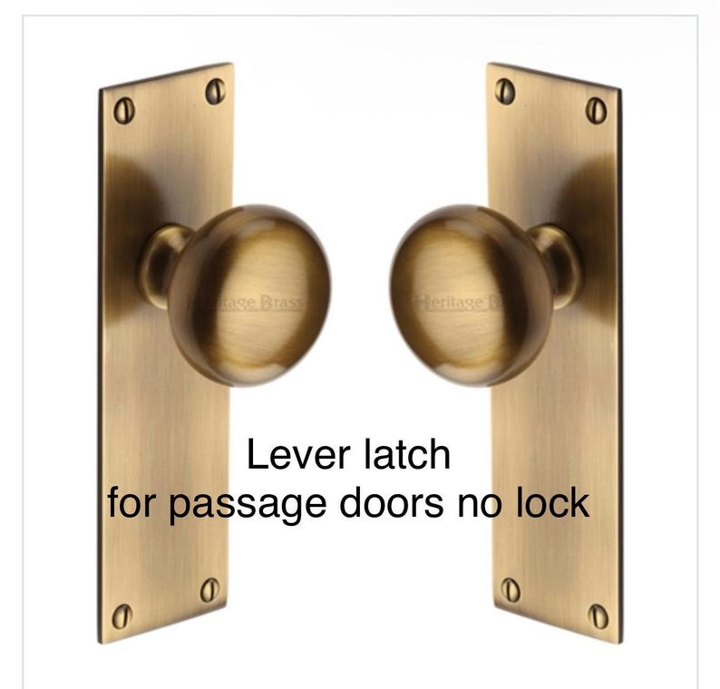 Peut inclure: Deux loquets &agrave; levier en laiton antique pour portes de passage, chacun avec un bouton rond et une plaque arri&egrave;re rectangulaire. Le texte "Lever latch for passage doors no lock" est imprim&eacute;.