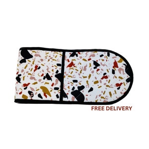 May include: An oven mitt featuring a terrazzo design in white, black, gold, red, and pink. It has a black border and a quilted pattern. The text "FREE DELIVERY" is printed in red on the bottom right.