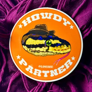 May include: A round orange sticker with a white border and white text that reads "Howdy Partner" in a retro font. A cartoon snake wearing a brown cowboy hat is in the center of the sticker. The snake is yellow, black, and green with a pink tongue.