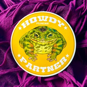 May include: A yellow circle sticker with the text "Howdy Partner" in a retro font. A cartoon frog wearing a cowboy hat is in the center of the sticker.