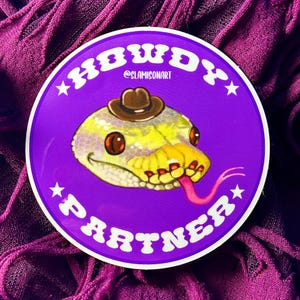 May include: A purple and white sticker with the text "Howdy Partner" in a retro font. A cartoon snake wearing a cowboy hat is featured in the center of the sticker.