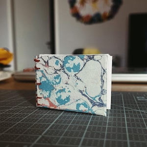 Pode incluir: Um pequeno caderno retangular com uma capa marmorizada apresentando padrões de redemoinho em azul, branco e castanho-avermelhado. O livro é encadernado no lado esquerdo com costura de fio vermelho visível e as páginas brancas são visíveis.