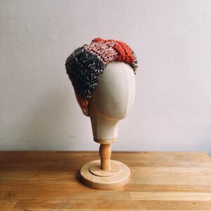 Puede incluir: Una diadema de ganchillo hecha a mano, multicolor, exhibida en una cabeza de maniquí. La diadema presenta una mezcla de hilo gris oscuro, verde, naranja y rosa. La cabeza está montada sobre un soporte de madera, sobre un fondo neutro.