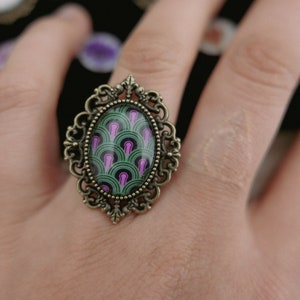 Könnte beinhalten: Ein Ring im Vintage-Stil mit einem ovalen grünen und violetten Kamee, der in einen verzierten Bronze-Rahmen gefasst ist.