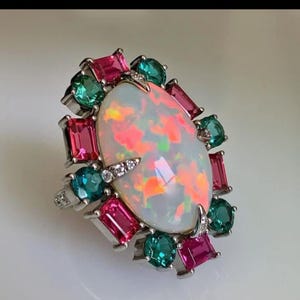 Puede incluir: Anillo de plata con una gran ópalo ovalado con vibrantes tonos rosa y naranja. El ópalo está rodeado de piedras preciosas verde esmeralda y magenta, con pequeñas piedras claras. Una pieza de joyería llamativa.