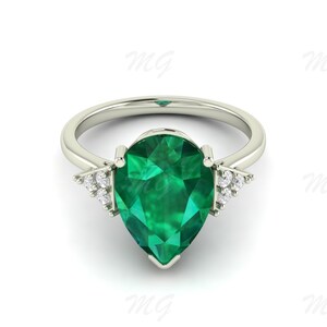 Puede incluir: Un anillo plateado con una gran piedra preciosa verde esmeralda en forma de pera. Pequeñas piedras claras están engastadas a ambos lados de la piedra principal. El anillo tiene un diseño simple y elegante, ideal para una ocasión especial.