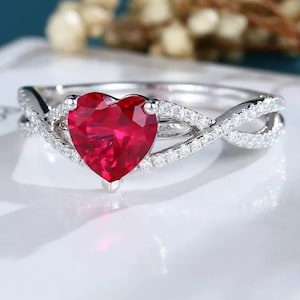 Puede incluir: Un anillo de plata con una piedra preciosa de rubí en forma de corazón rodeada de diamantes más pequeños. La banda tiene un diseño retorcido con diamantes engastados a lo largo de los bordes.