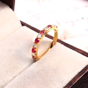 Puede incluir: Un anillo de banda de oro con piedras preciosas rojas y blancas alternas. El anillo está en una caja de anillo de terciopelo.