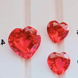 Puede incluir: Tres piedras preciosas rojas talladas en forma de corazón de diferentes tamaños dispuestas sobre una superficie blanca con marcas numéricas negras parciales visibles a los lados, mostrando un corte brillante y un color carmesí vibrante.