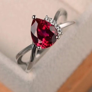 Puede incluir: Un anillo de plata con una piedra roja en forma de pera y piedras transparentes más pequeñas en la banda.