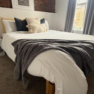 May include: A bed with a white duvet cover and a grey knitted throw. Decorative pillows in yellow, navy, and cream colours are arranged on the bed. A framed picture hangs above the bed, set against a brick wall.