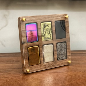 May include: A wooden display case with a clear acrylic front, showcasing six different Zippo lighters. The lighters are arranged in a grid pattern, with each lighter having a unique design. The case is made of dark wood with brass accents and the word "Zippo" is engraved on the bottom.