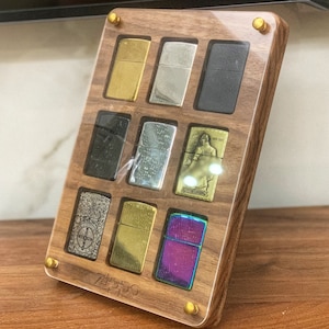 Pode incluir: Uma vitrine de madeira com nove isqueiros de diferentes cores e padrões. A vitrine é feita de madeira escura com uma tampa de acrílico transparente. Os isqueiros estão dispostos em uma grade. A vitrine tem uma etiqueta de metal dourado com a palavra "Zippo" impressa nela.