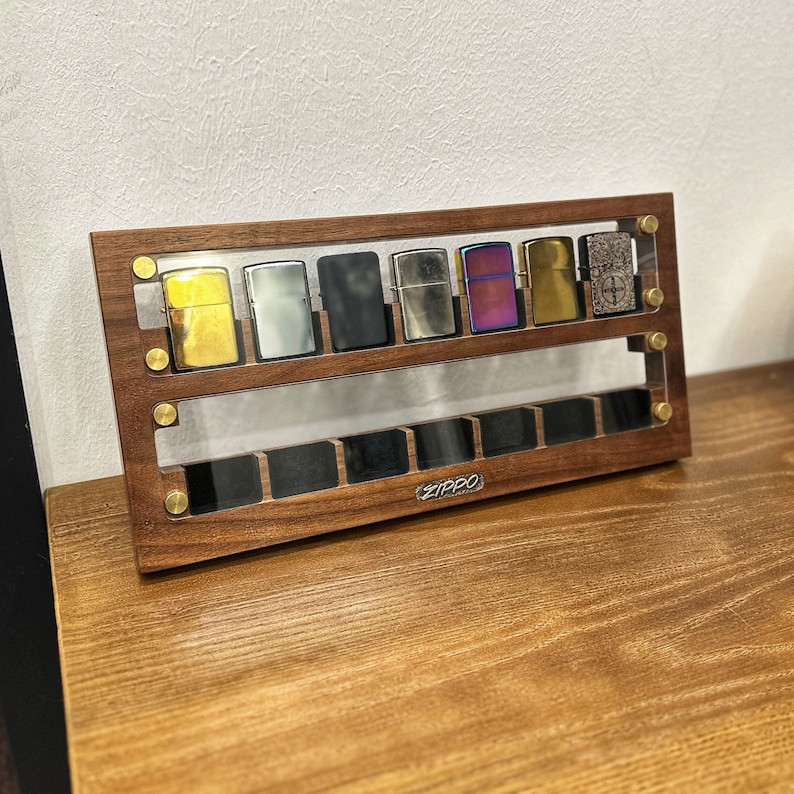 May include: A wooden display case with a clear acrylic front, showcasing a collection of eight silver and gold lighters. The case has a dark wood finish and the word "ZIPPO" is engraved on the front.