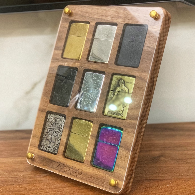 May include: A wooden display case with nine different colored and patterned lighters. The case is made of dark wood with a clear acrylic top. The lighters are arranged in a grid pattern. The case has a gold metal label with the word "Zippo" printed on it.