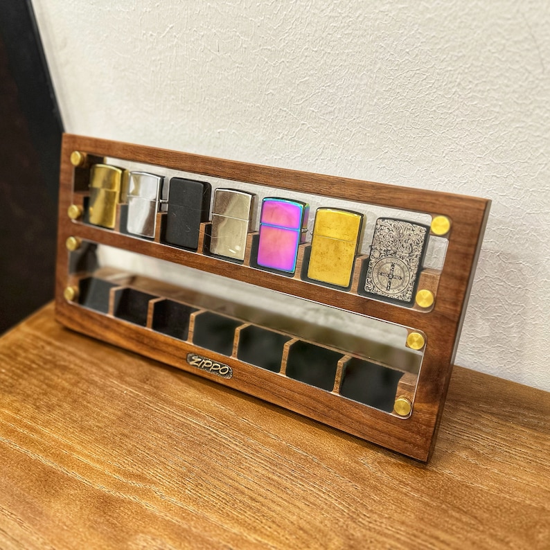 May include: A wooden display case with a clear acrylic front, showcasing a collection of eight silver and gold lighters. The lighters are arranged in a row, with the brand name "ZIPPO" visible on the case.