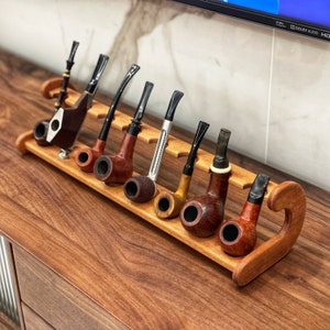 May include: A wooden stand with eight tobacco pipes. The pipes are made of different materials, including wood, metal, and ceramic. The stand is a dark brown color and has a curved design.
