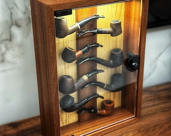 Puede incluir: Una vitrina de madera con frente de vidrio, que muestra una colección de seis pipas de tabaco. Las pipas están dispuestas verticalmente, con las boquillas mirando al espectador. La vitrina tiene un acabado de madera oscura y un cierre de latón.