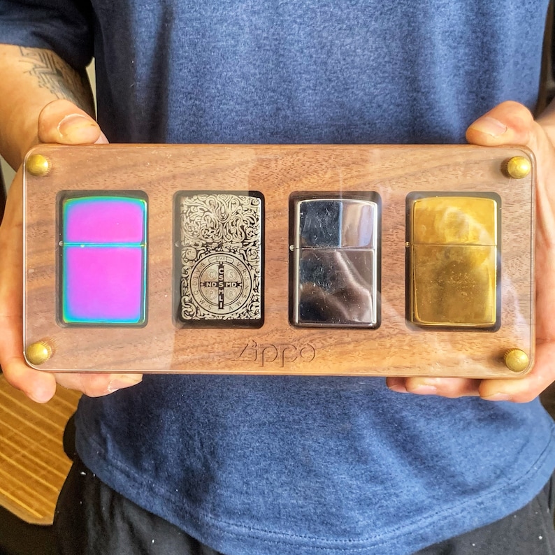 May include: A wooden box with a glass top displaying four different Zippo lighters. The lighters are in a variety of finishes, including chrome, gold, and a rainbow iridescent finish. The box is labeled "Zippo" on the front.