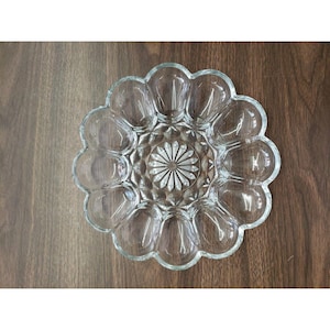 May include: Clear glass egg serving dish with a flower-like design. The dish has 12 egg-shaped compartments around a central starburst pattern. The edges of the dish are scalloped. The dish is sitting on a dark brown wooden surface.