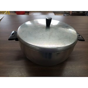 May include: A vintage aluminum cooking pot with a lid. The pot has a rounded shape with two black handles and a black handle on the lid. The pot is sitting on a wooden surface.